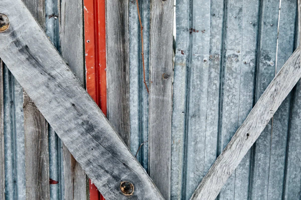 Weathered vertical wooden slats with peeling gray paint, crossed by a diagonal weathered board and a narrow red strip visible behind.
