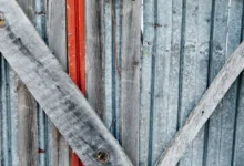 Weathered vertical wooden slats with peeling gray paint, crossed by a diagonal weathered board and a narrow red strip visible behind.
