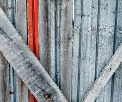Weathered vertical wooden slats with peeling gray paint, crossed by a diagonal weathered board and a narrow red strip visible behind.