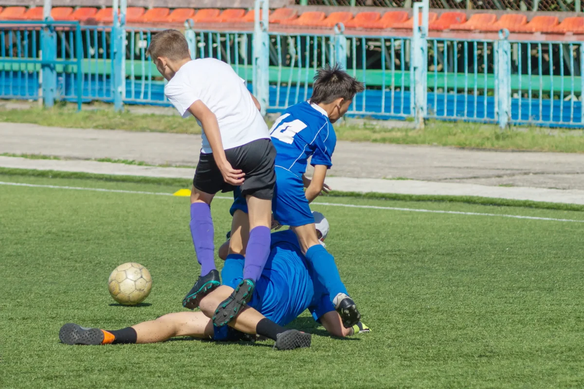 Youth soccer game: several players in white and blue battle for the ball on a sunny grass field, one player sliding in.
