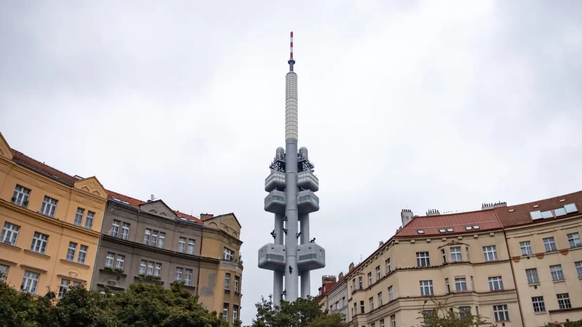 Tall communications tower rises above surrounding city buildings under a cloudy sky, centered in the frame.