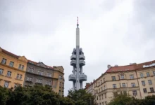 Tall communications tower rises above surrounding city buildings under a cloudy sky, centered in the frame.