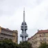 Tall communications tower rises above surrounding city buildings under a cloudy sky, centered in the frame.