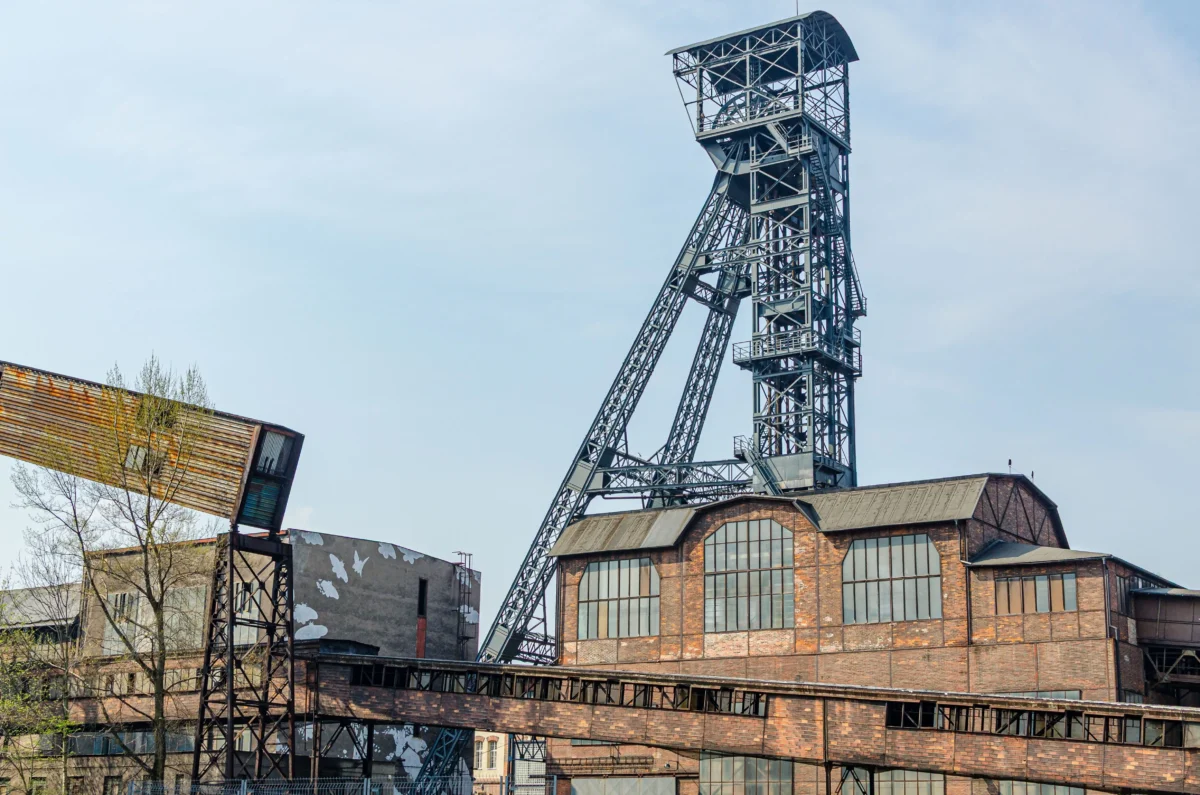 shutterstock_2692845229 1 | TVGURU.cz Tall steel lattice mining tower rises beside brick factory buildings under a blue sky, in an industrial site setting.