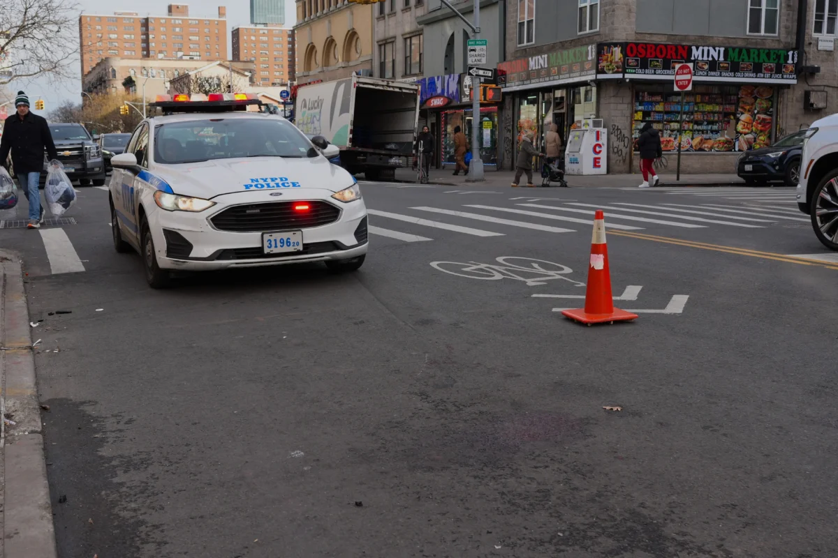 shutterstock_2727104759 1 | TVGURU.cz NYPD police car parked on a city street with a red light, an orange traffic cone on a crosswalk, and pedestrians nearby near storefronts.