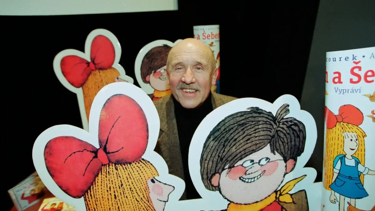 Smiling older man with a mustache posing behind large cartoon cutouts of children with big red bows at a book display.