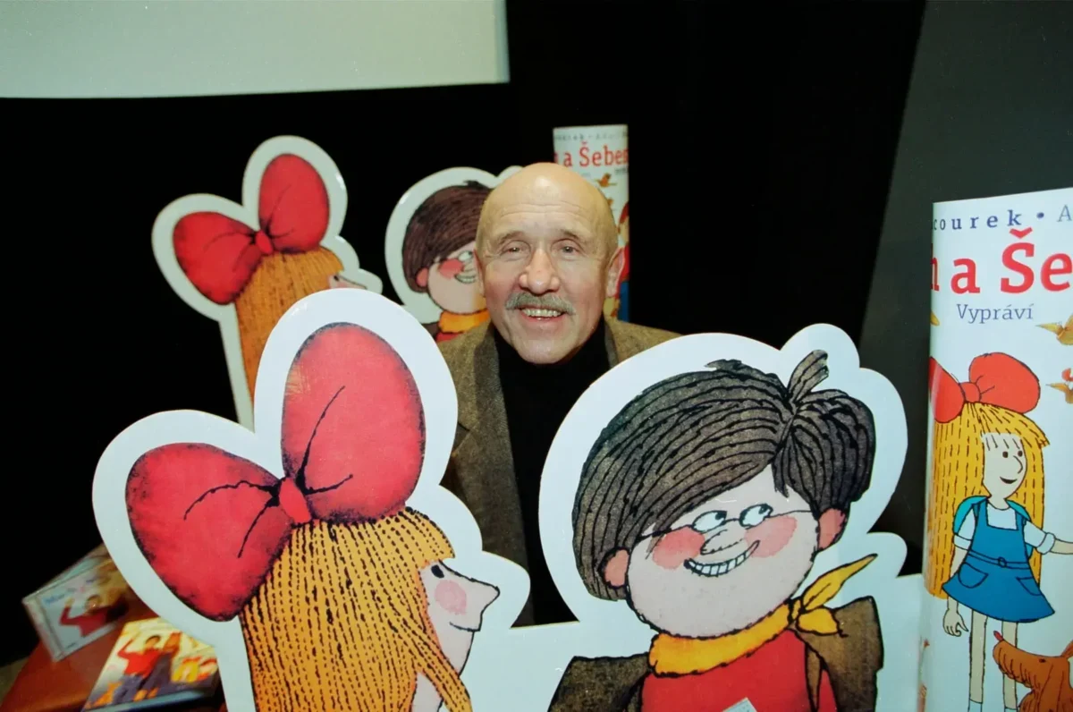 Smiling older man with a mustache posing behind large cartoon cutouts of children with big red bows at a book display.