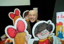 Smiling older man with a mustache posing behind large cartoon cutouts of children with big red bows at a book display.