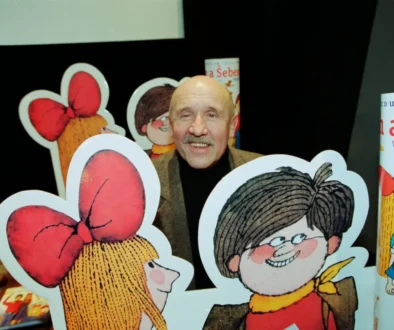 Smiling older man with a mustache posing behind large cartoon cutouts of children with big red bows at a book display.
