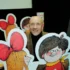 Smiling older man with a mustache posing behind large cartoon cutouts of children with big red bows at a book display.