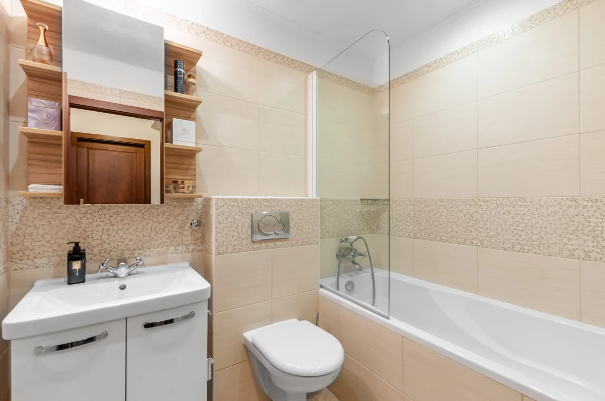 shutterstock_1922430992 1 | TVGURU.cz Modern beige-tiled bathroom with a sink, toilet, and glass-enclosed bathtub/shower combo, plus wooden shelving units along the wall.