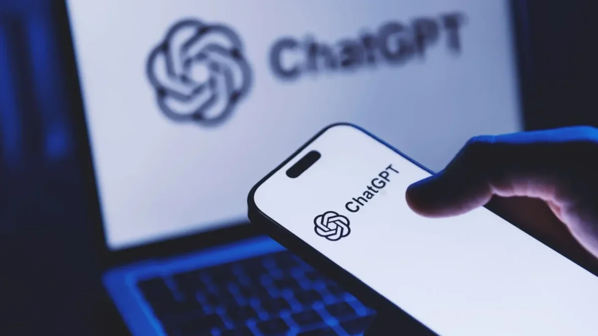 Close-up of a smartphone displaying the ChatGPT logo, with a laptop screen showing ChatGPT in the blurred background.