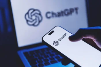 Close-up of a smartphone displaying the ChatGPT logo, with a laptop screen showing ChatGPT in the blurred background.