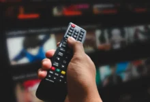 Close-up of a hand holding a TV remote, aimed at a blurry television screen in the background.