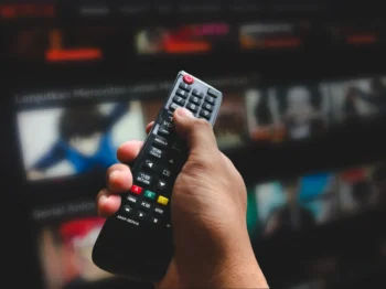 Close-up of a hand holding a TV remote, aimed at a blurry television screen in the background.