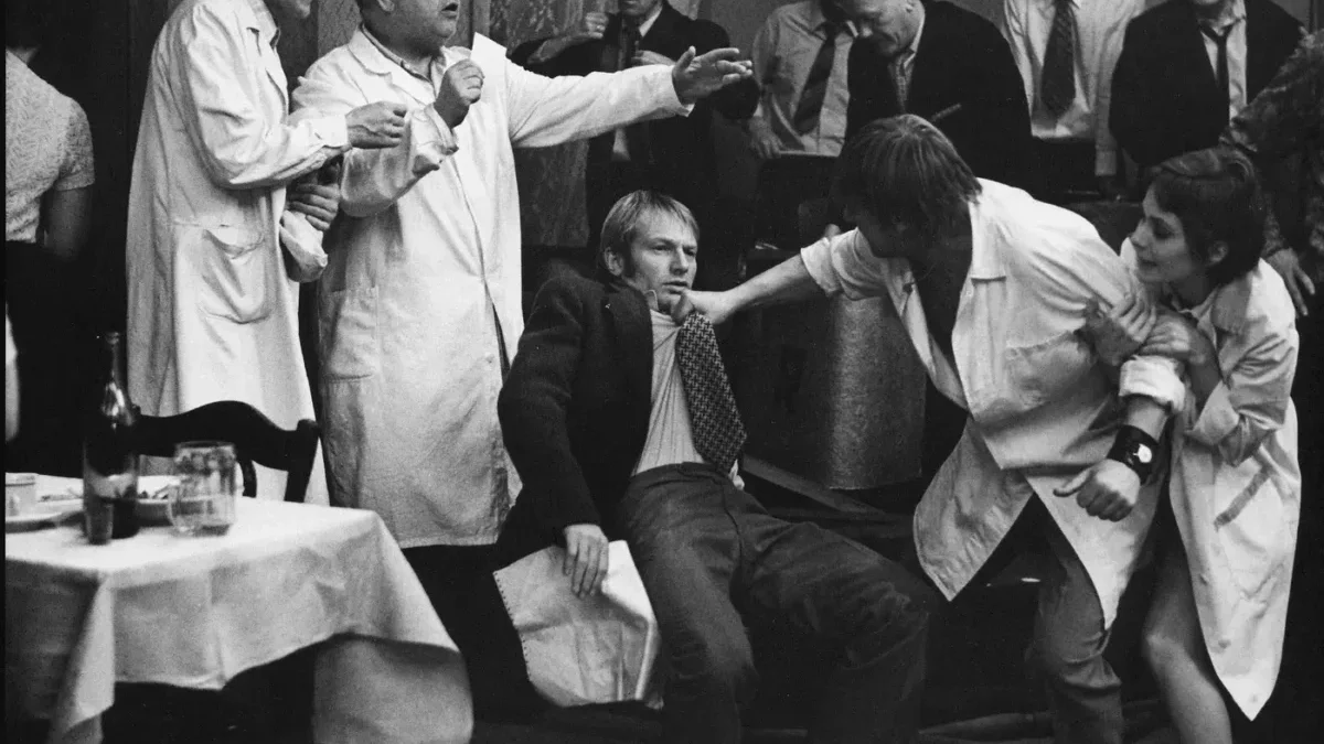 A man in a suit is restrained by two people in lab coats while others watch a tense scene at a social gathering.