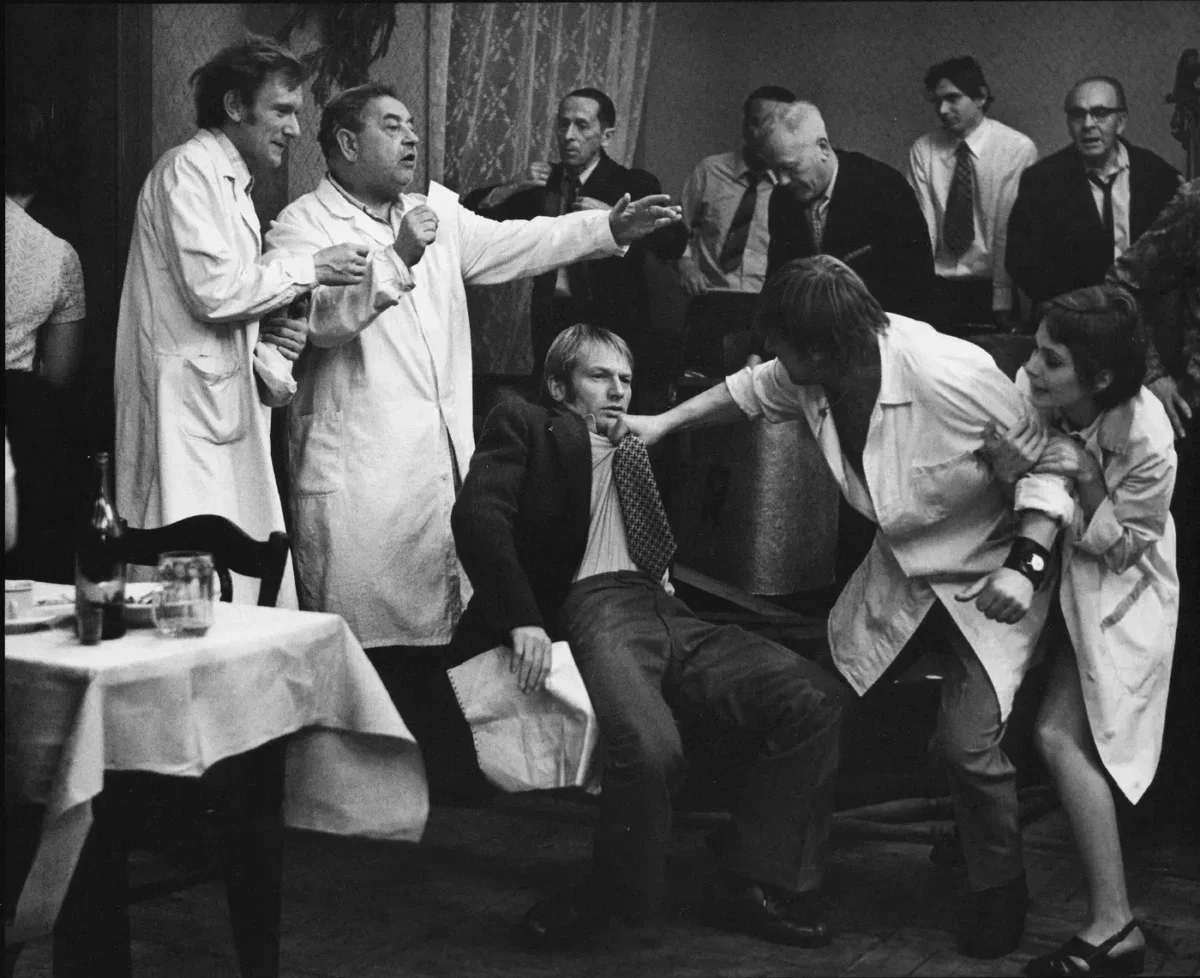 A man in a suit is restrained by two people in lab coats while others watch a tense scene at a social gathering.