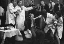 A man in a suit is restrained by two people in lab coats while others watch a tense scene at a social gathering.