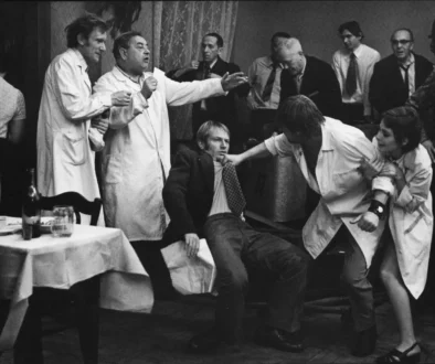 A man in a suit is restrained by two people in lab coats while others watch a tense scene at a social gathering.