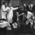 A man in a suit is restrained by two people in lab coats while others watch a tense scene at a social gathering.