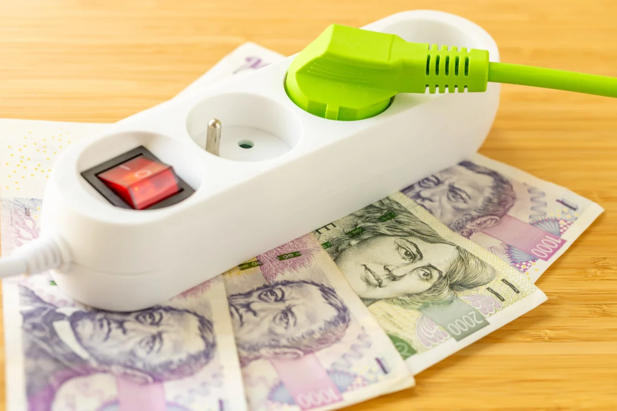 shutterstock_2643708313 1 | TVGURU.cz White power strip with a bright green plug inserted, resting on scattered banknotes on a wooden table.