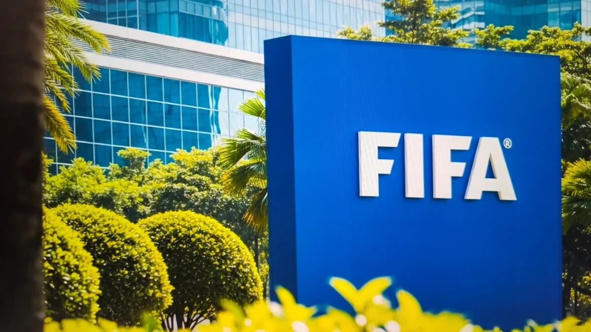 Blue FIFA sign outdoors with white letters, surrounded by green shrubs and a glass-front building in the background.