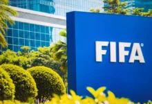 Blue FIFA sign outdoors with white letters, surrounded by green shrubs and a glass-front building in the background.