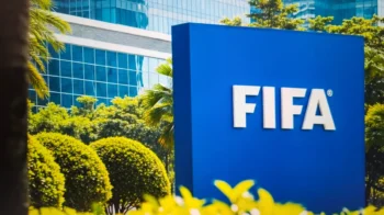 Blue FIFA sign outdoors with white letters, surrounded by green shrubs and a glass-front building in the background.