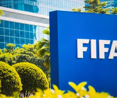 Blue FIFA sign outdoors with white letters, surrounded by green shrubs and a glass-front building in the background.
