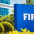 Blue FIFA sign outdoors with white letters, surrounded by green shrubs and a glass-front building in the background.