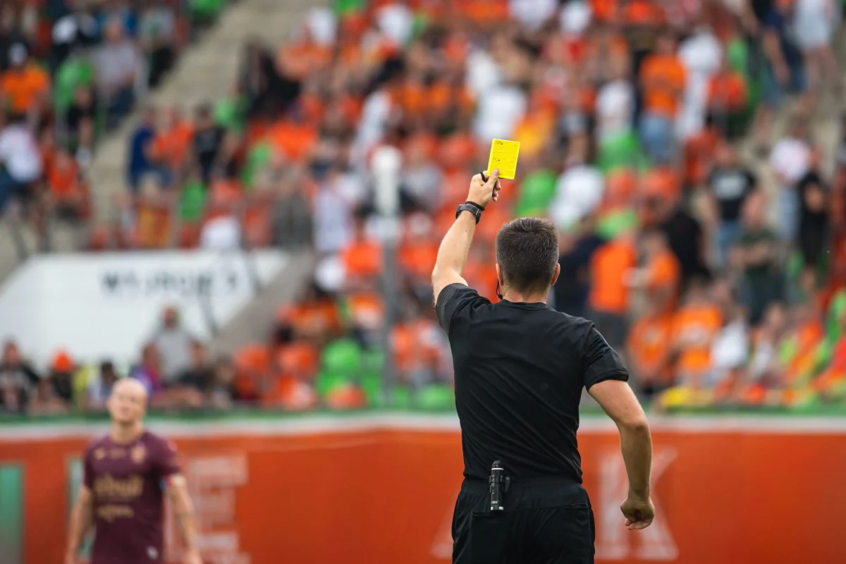 shutterstock_2694301153 1 | TVGURU.cz Soccer referee in black raises a yellow card to caution a player, crowd blurred in the stadium behind.