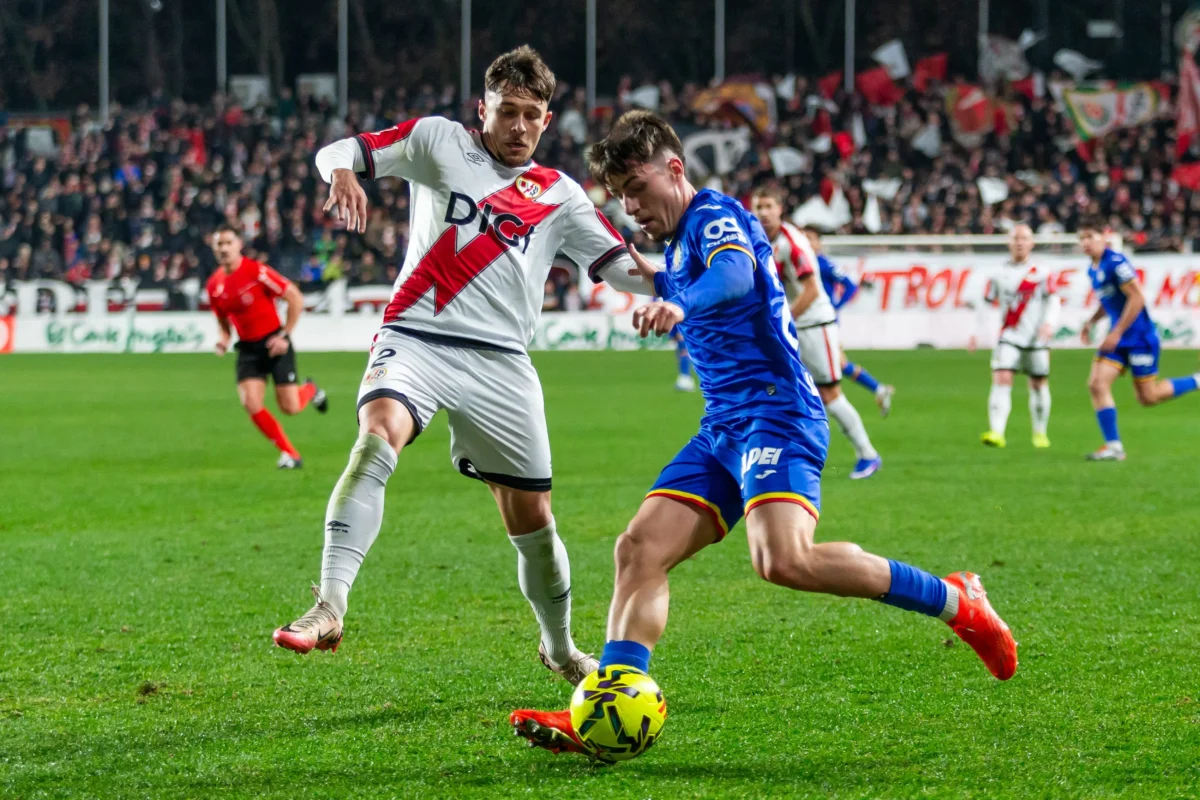 shutterstock_2721572489 1 | TVGURU.cz Two soccer players, one in white with a red diagonal stripe and one in blue, chase a neon yellow ball on a green pitch as fans watch.