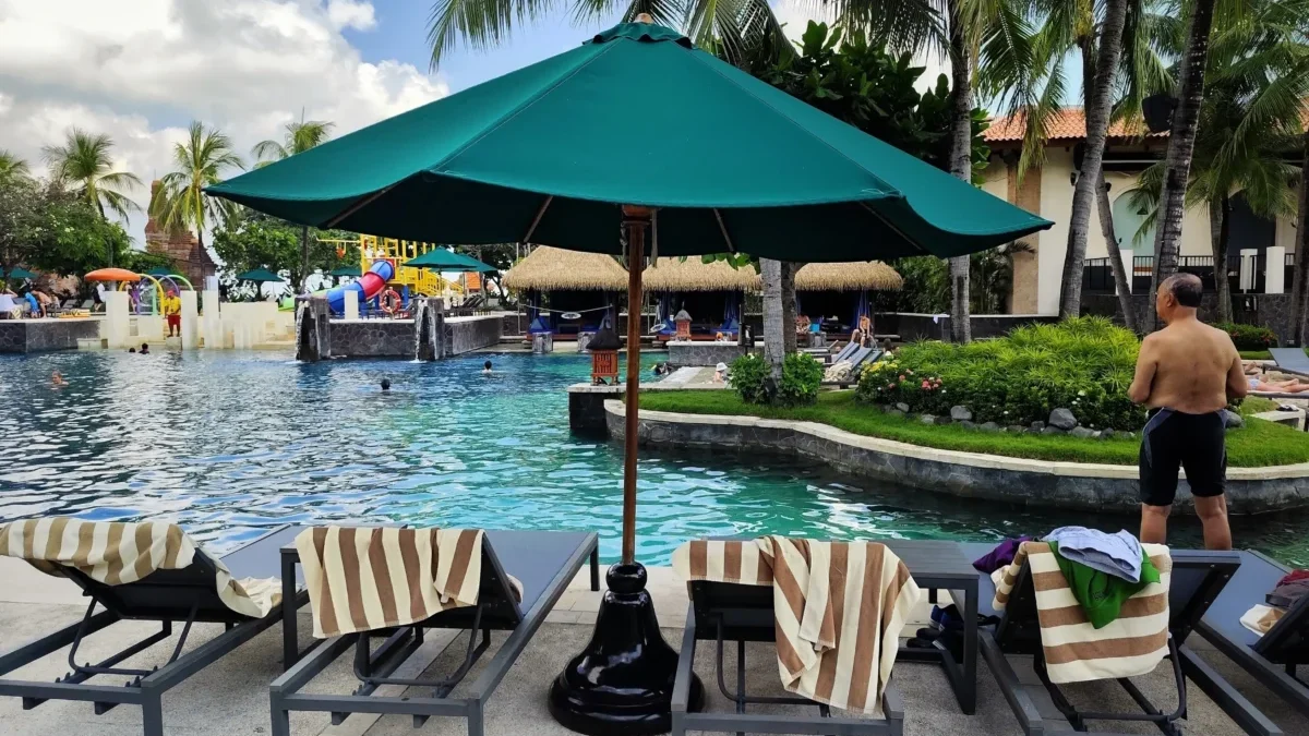 Resort pool area with striped lounge chairs under a green umbrella, palm trees, and people relaxing by the water.