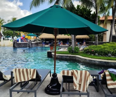 Resort pool area with striped lounge chairs under a green umbrella, palm trees, and people relaxing by the water.