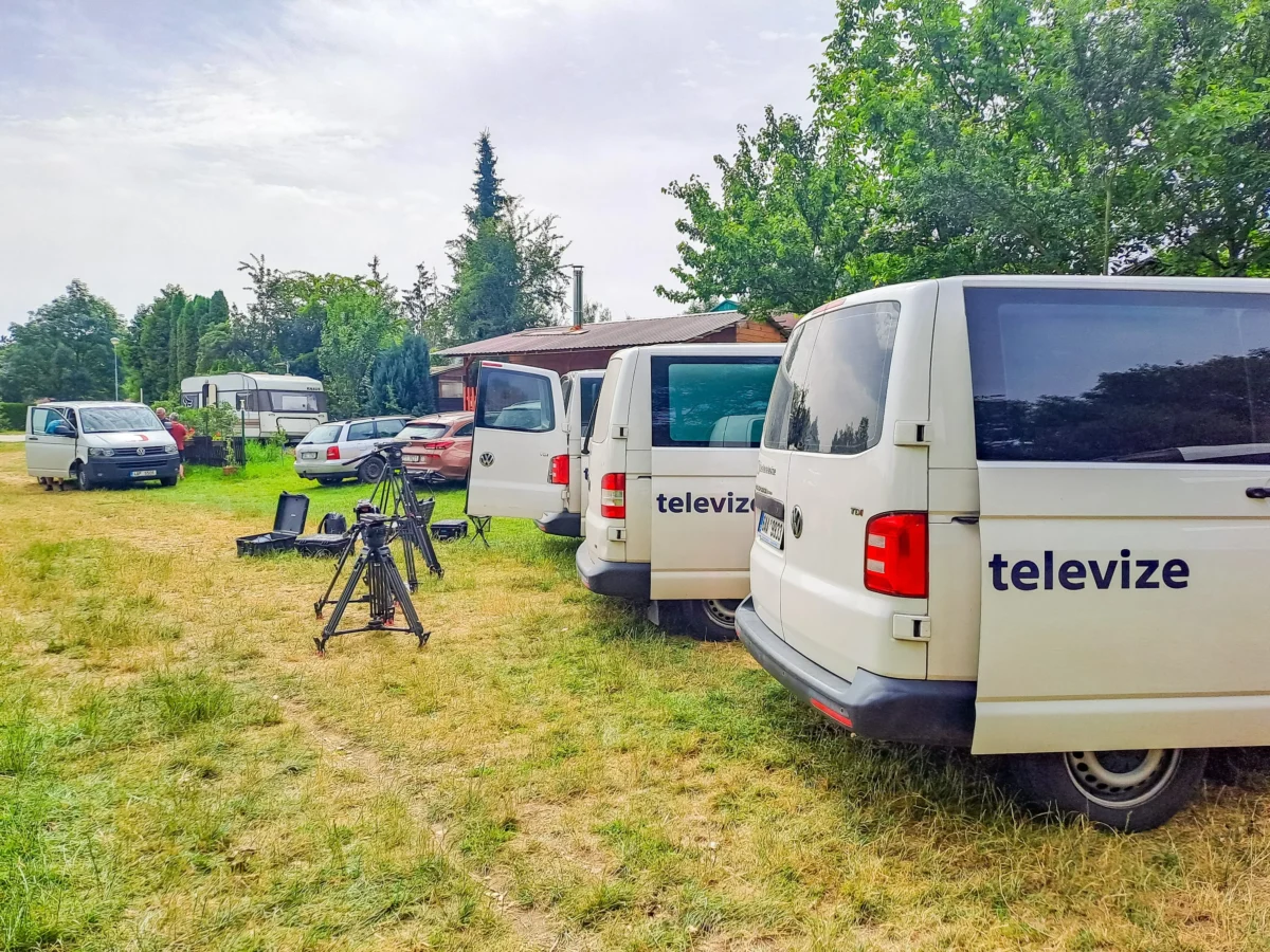 shutterstock_2481637251 1 | TVGURU.cz White TV production vans parked in a grassy lot with camera gear and tripods in the foreground on a sunny day.