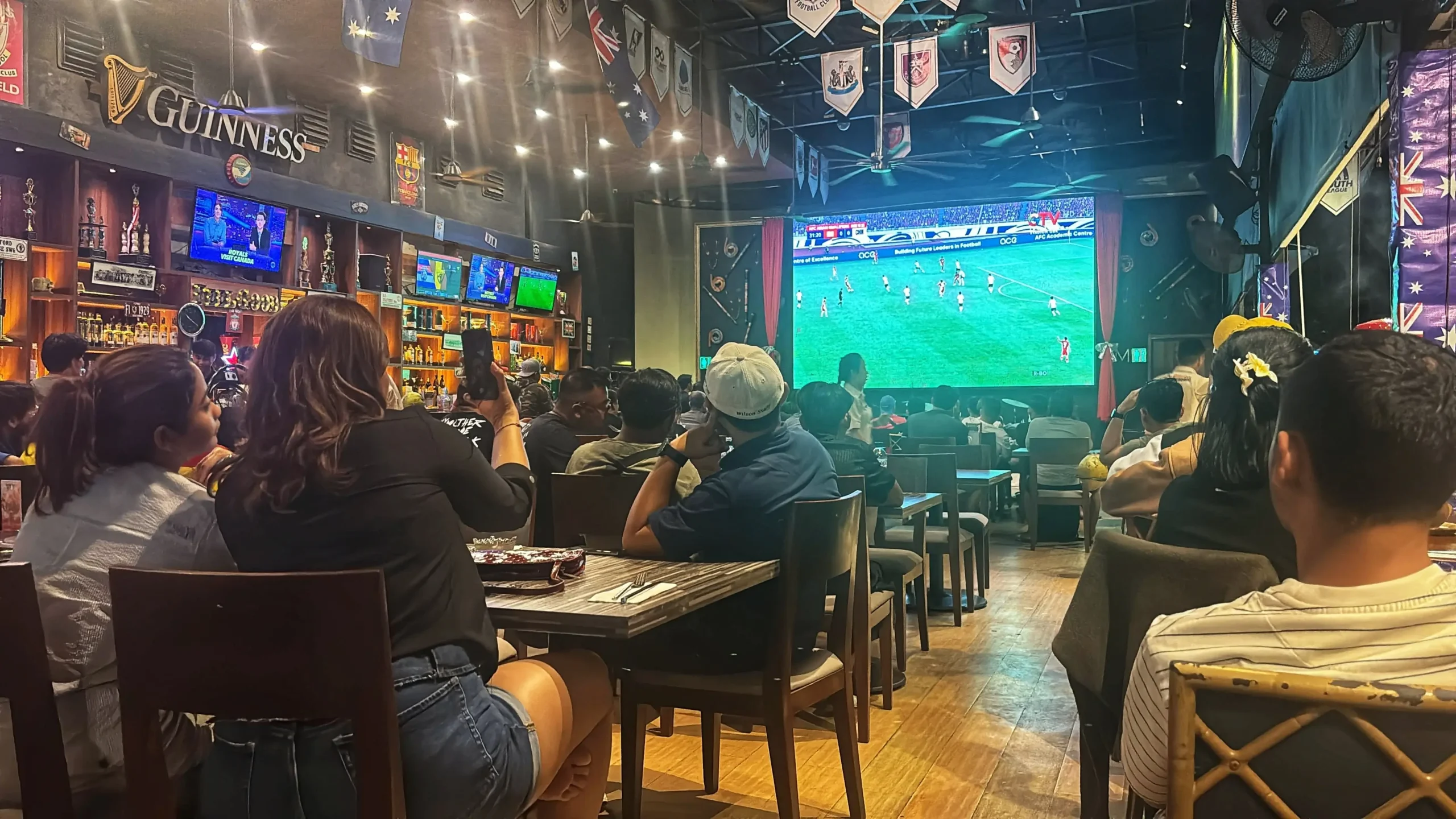 Lidé sledují fotbal na projektoru v hospodě.
