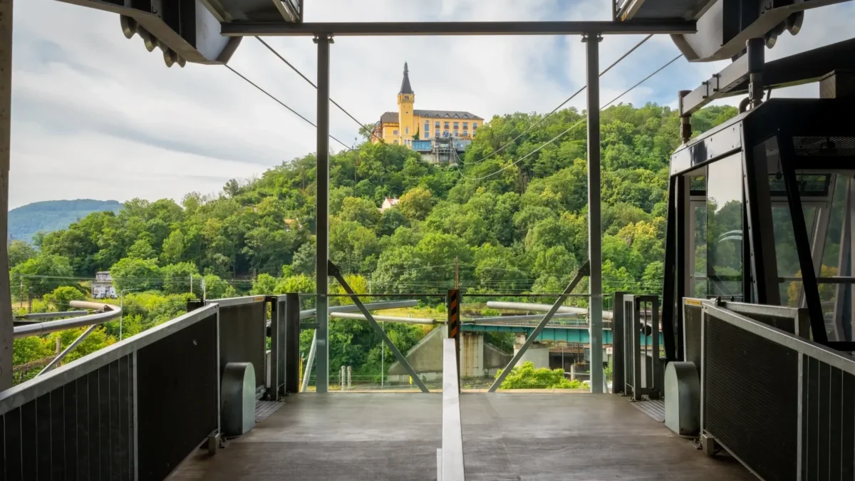 View from a tram platform toward a yellow hilltop building surrounded by green trees, with metal framework and rails in the foreground.