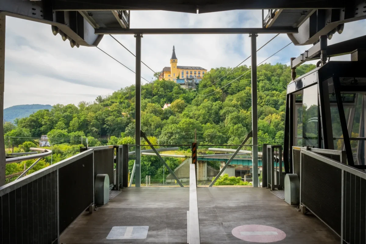 View from a tram platform toward a yellow hilltop building surrounded by green trees, with metal framework and rails in the foreground.