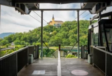 View from a tram platform toward a yellow hilltop building surrounded by green trees, with metal framework and rails in the foreground.
