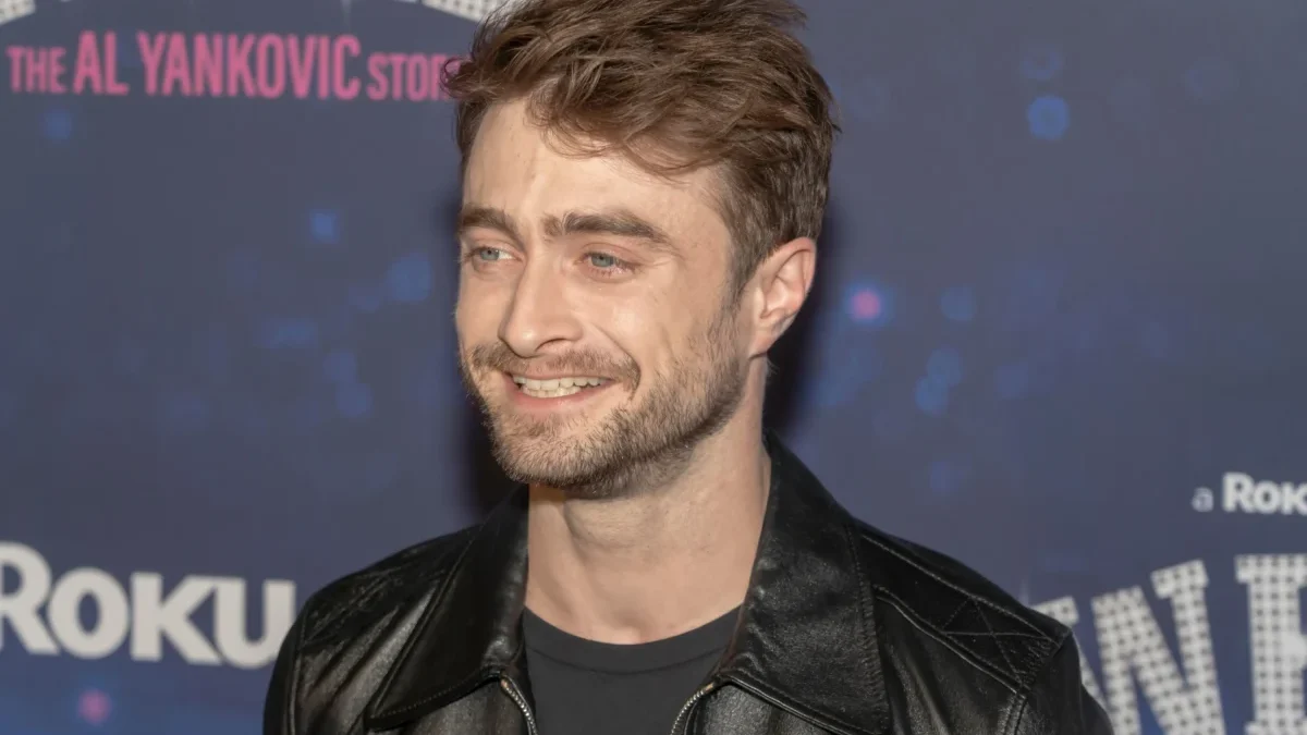 Man with short brown hair and stubble wearing a black leather jacket, smiling at a backdrop-covered event wall.