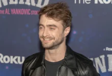 Man with short brown hair and stubble wearing a black leather jacket, smiling at a backdrop-covered event wall.