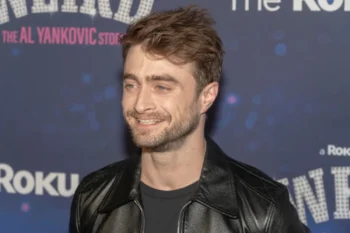 Man with short brown hair and stubble wearing a black leather jacket, smiling at a backdrop-covered event wall.