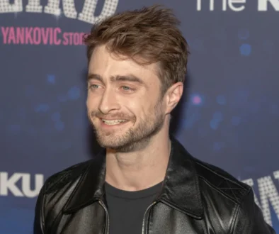 Man with short brown hair and stubble wearing a black leather jacket, smiling at a backdrop-covered event wall.