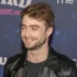 Man with short brown hair and stubble wearing a black leather jacket, smiling at a backdrop-covered event wall.