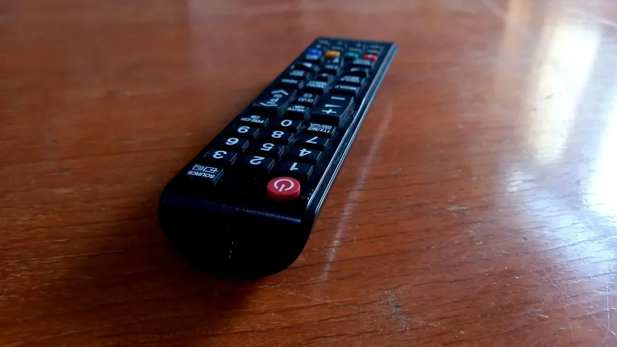 Black TV remote lying on a wooden table, angled to the left with a red power button visible at the bottom corner.