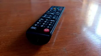 Black TV remote lying on a wooden table, angled to the left with a red power button visible at the bottom corner.