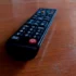 Black TV remote lying on a wooden table, angled to the left with a red power button visible at the bottom corner.