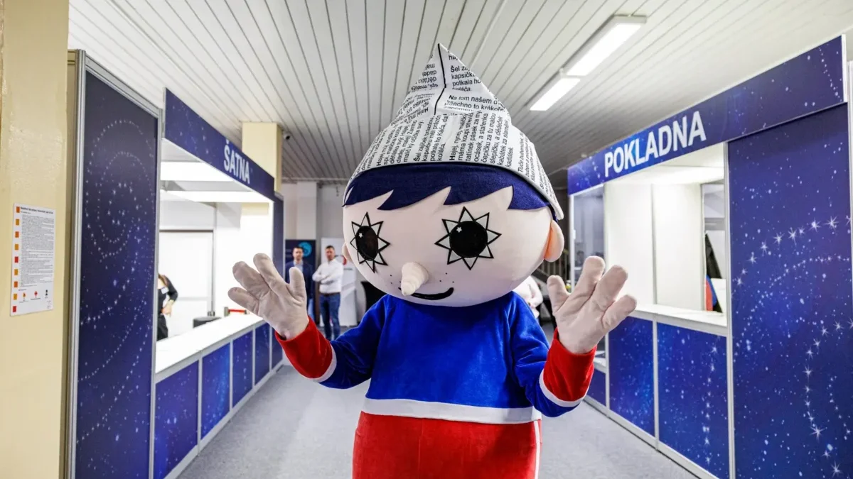 Mascot with a newspaper cone hat stands in a blue service hall, waving at the camera.