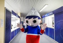 Mascot with a newspaper cone hat stands in a blue service hall, waving at the camera.