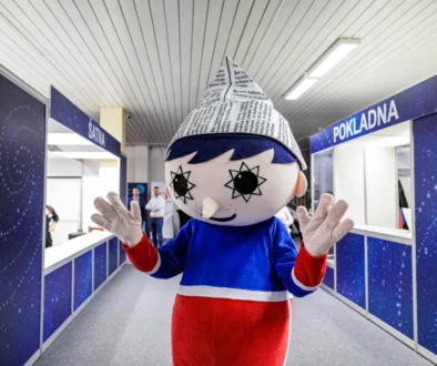 Mascot with a newspaper cone hat stands in a blue service hall, waving at the camera.